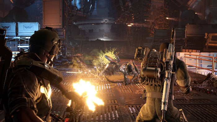 A dramatic action scene from a video game showing two heavily armored soldiers in a futuristic industrial setting, with one firing a bright flame weapon and the other holding a rifle, while a third figure lies defeated in the center.