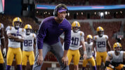 Football coach in a purple jersey with players in yellow and gray uniforms on a field.