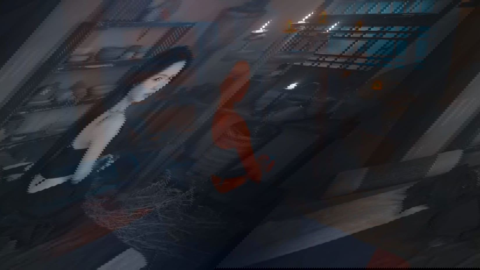 Woman in a black outfit sitting on a bed in a dimly lit room with candles and shelves.