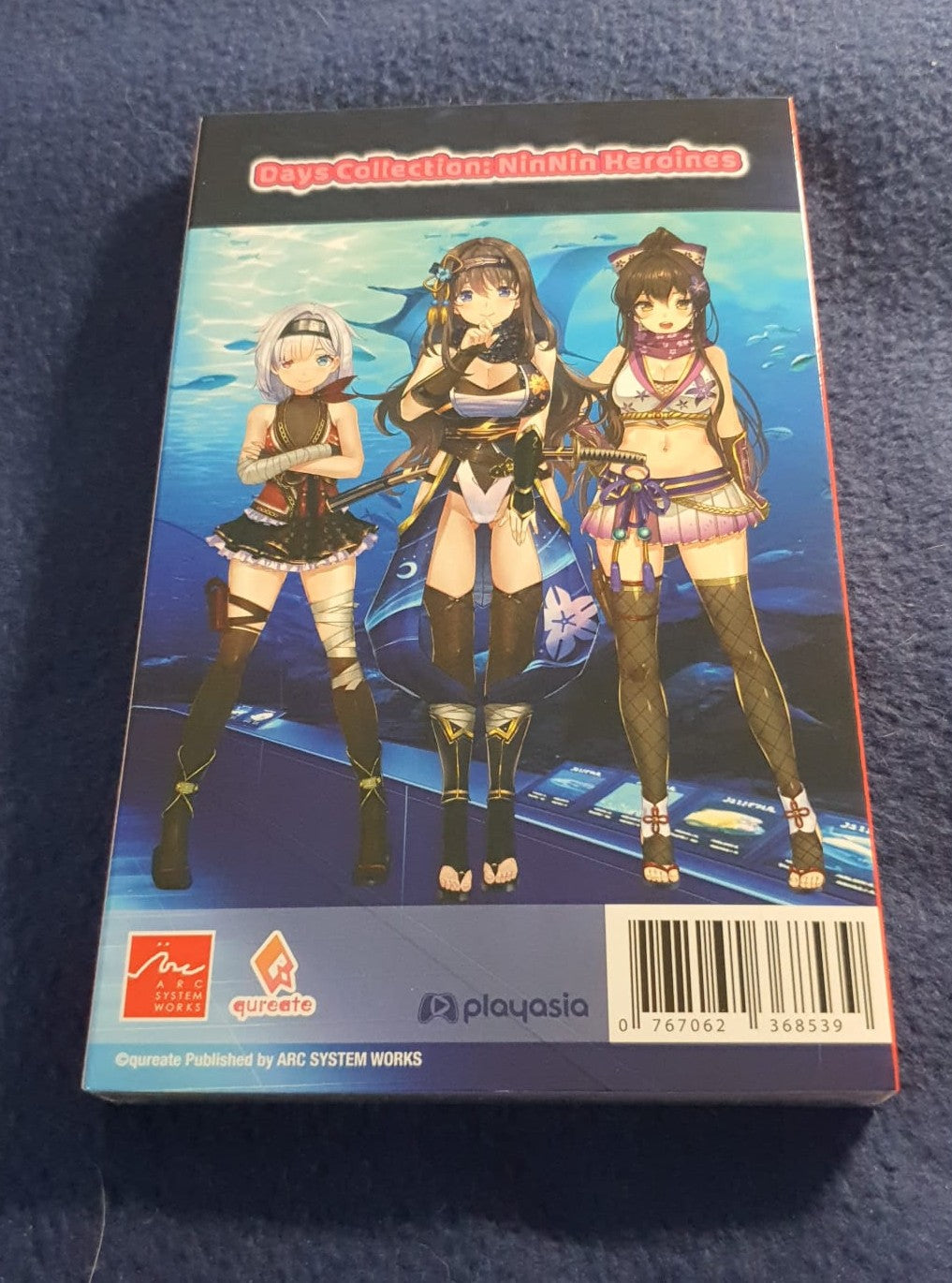 A boxed collectible figure set photographed from above on a dark blue textured fabric surface. The front of the box features anime-style female characters against a blue underwater scene, with visible publisher marks including Quatro Sogei and Playasia.