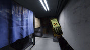 A hand holding a yellow handheld electronic device with a digital display and keypad, pointing it down a narrow industrial corridor with metal mesh gates and a blue fabric cover on the left.