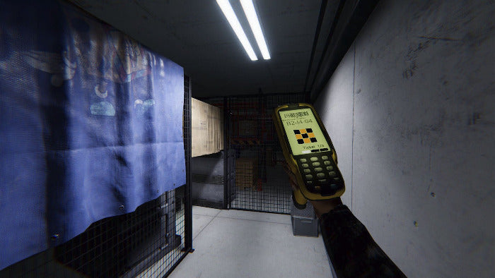 A hand holding a yellow handheld electronic device with a digital display and keypad, pointing it down a narrow industrial corridor with metal mesh gates and a blue fabric cover on the left.