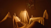 A moody, dimly lit scene showing draped fabric-covered forms in a dark room, with candles placed on top of the drapes and a small framed artwork hanging on the wall.