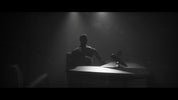 A dark, moody scene with a silhouetted seated figure at a table under spotlights, with a microphone in front. The image is heavily shadowed and low-key, creating a dramatic, cinematic look.