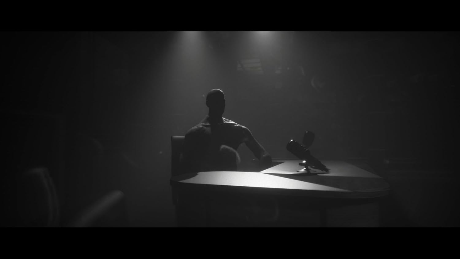A dark, moody scene with a silhouetted seated figure at a table under spotlights, with a microphone in front. The image is heavily shadowed and low-key, creating a dramatic, cinematic look.
